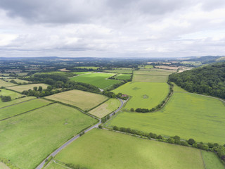 Aerial view of green wheat farm fields, with a crossing country roads, woodland on the hill, in an English countryside, on a cloudy summer day .