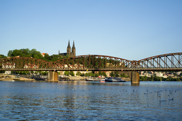 Railway bridge at Vyton under Vysehrad, Prague, Czech Republic / Czechia. Vltava river, embankment with unidentifiable people,. Building of church and cathedral on the top of hill. Sunny sunset light