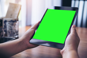 Mockup image of hands holding black tablet pc with white blank green screen on wooden table background in coffee shop