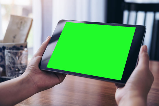 Mockup image of hands holding black tablet pc with white blank green screen on wooden table background in coffee shop