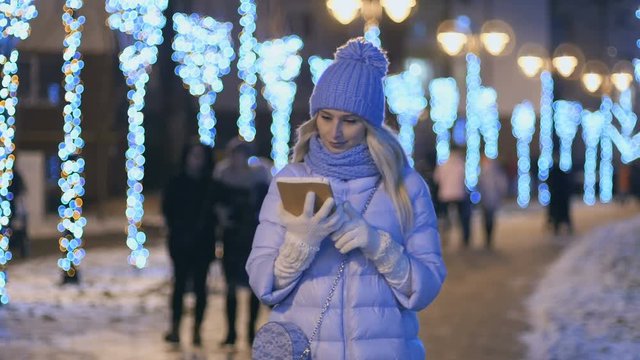 Beautiful blonde in winter clothes uses a tablet at night lights background