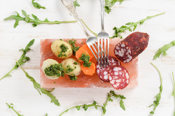 Salami, potatoes, carrots and fresh arugula with green chimichurri sauce served on pink hymalayan salt block on white background