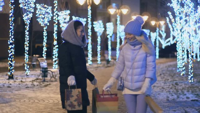 Two girls with packages shows each other gifts and happy walks at night city