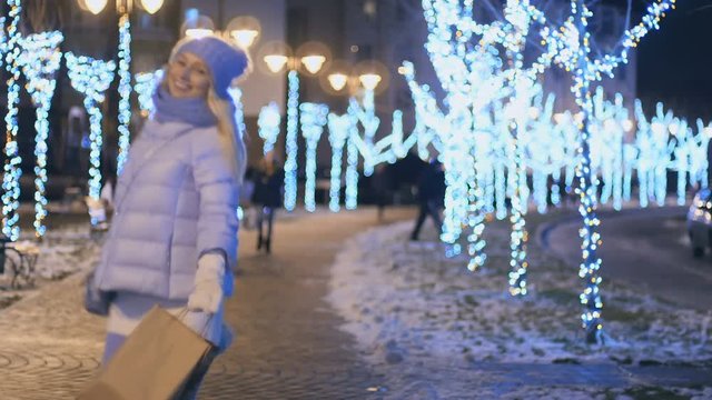 Happy blonde with packages walks at lights night city at winter