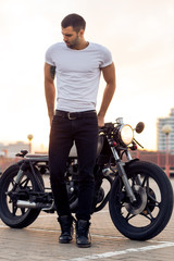 Sporty biker handsome rider man in white blank t-shirt walk away from classic style cafe racer motorbike at sunset. Vintage bike custom made in garage. Brutal urban lifestyle. Outdoor portrait. - 165006029