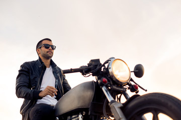 Handsome rider male with beard and mustache in black biker jacket, white shirt and fashion sunglasses smoke cigaret and sit on classic style cafe racer motorbike at sunset. Brutal fun urban lifestyle.