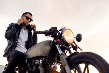 Fototapeta na wymiar Handsome rider guy with beard and mustache in black fashion sunglasses smoking cigaret and correct biker jacket sit on classic style cafe racer motorbike at sunset. Brutal fun urban lifestyle.