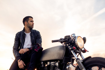 Handsome rider boy with beard and mustache in black leather biker jacket sit on classic style cafe racer motorbike on rooftop at sunset. Bike custom made in vintage garage. Brutal fun urban lifestyle.