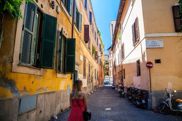 Fototapeta na wymiar Girl in red dress walking on Rome street. Trastevere, Rome, Italy. Young girl. Old street in Rome. Pretty girl around the corner. Rome architecture. Blonde hair girl