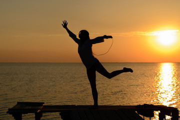 A girl in lingerie and shirt listening music on headphones on a pier by the sea at sunset