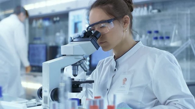 Medical Research Scientists putting Slides in Place and Looking at Samples Under Microscope. She Works in a Bright Modern Laboratory. Shot on RED EPIC-W 8K Helium Cinema Camera.