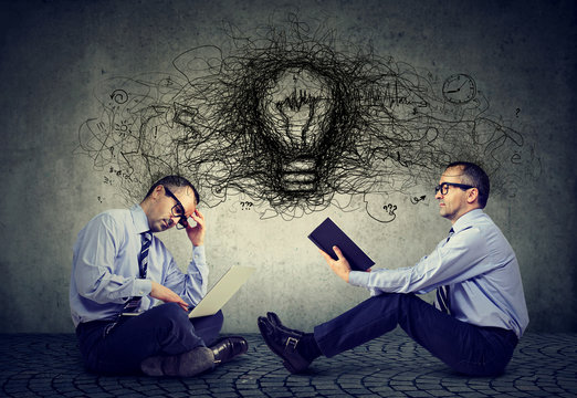 Businessmen sitting on a floor in office one using laptop another reading a book with light bulb and idea thoughts in-between  