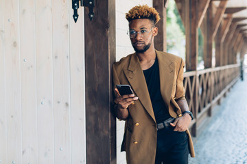 A portrait of African American man in jacket and glasses with smartphone standing near the wooden...