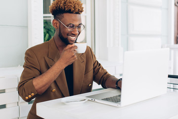 Portrait of an African American man in a jacket and glasses who drink coffee and work on a laptop