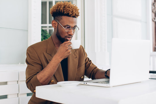 Portrait of an African American man in a jacket and glasses who drink coffee and work on a laptop
