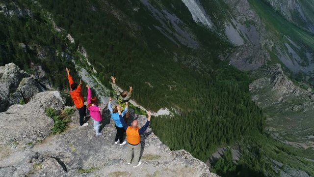 Climbers on summit. Happy people embracing raising hands up on pointed rocky cliff blue sky background aerial slow motion 4k
