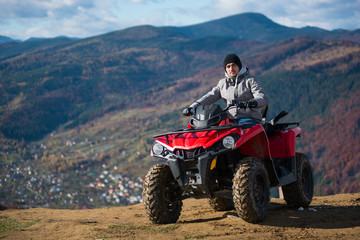 Fototapeta na wymiar Guy in winter clothes on a red quad bike on a mountain top looking at the camera. Blurred background of mountain scenery