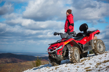 Male rider in a red winter clothes with red quad bike standing on a snowy mountain slope under the blue cloudy sky on a blurred background of mountains and the town in the valley with copy space