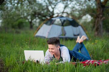 Obraz premium Young smiling woman lying on the grass working on her laptop while camping outdoors her tent on the background copyspace relaxing studying headphones technology gadget