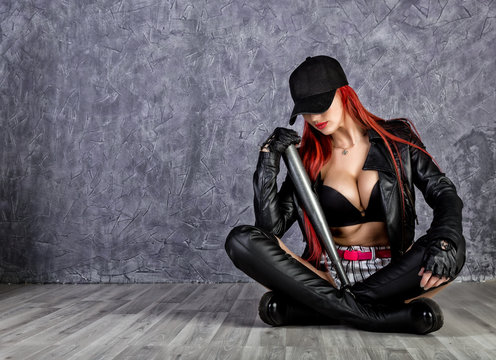 Stylish hipster woman with big tits in baseball cap and black jacket sitting on a floor, holding baseball bat.