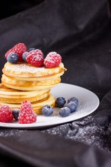 Stack of pancakes with raspberries and blueberries coated with powdered sugar  on white plate and black background