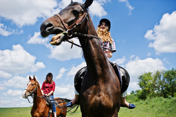 Tow young pretty girls riding a horses on a field at sunny day