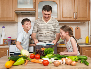 Father and two children reading cooking book and choice dishes. Happy family, girl and boy having fun with fruits and vegetables in home kitchen interior. Healthy food concept.