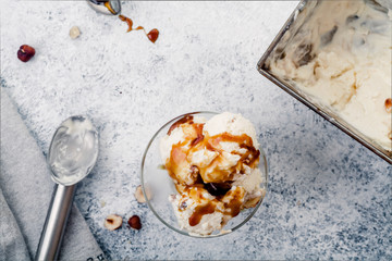 Caramel ice cream scooped in glass bowl from metallic container. Peanuts and chocolate used for decoration. Top view with copy space. Rustic textured background.