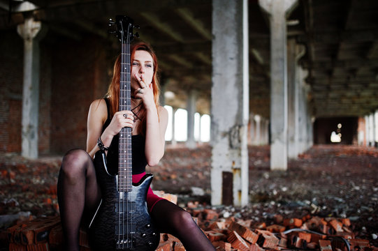 Red haired punk girl wear on black and red skirt, with bass guitar at abadoned place. Portrait of gothic woman musician.Smoking cigarette.