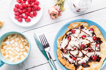 Homemade pancakes or Russian pancakes with chocolate sauce, whipped cream and raspberries on a plate on a white wooden background.