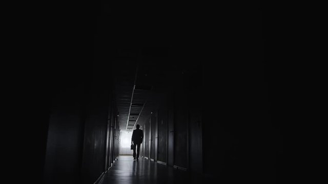PAN with wide shot of silhouette of confident man with briefcase walking along dark corridor in office centre 