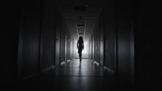 Tilt up of silhouette of unrecognizable businesswoman walking elegantly along dark corridor towards camera 