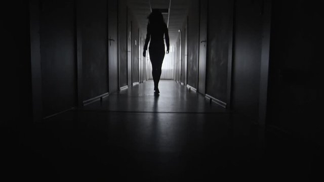 Tilt up of silhouette of unrecognizable businesswoman walking along dark corridor towards only window 
