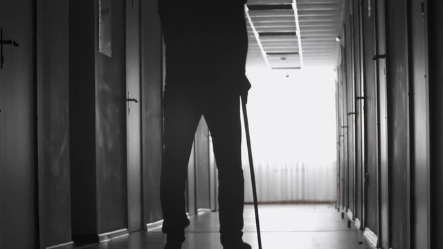 Tilt up of silhouette of man with limp walking along dark corridor using stick 