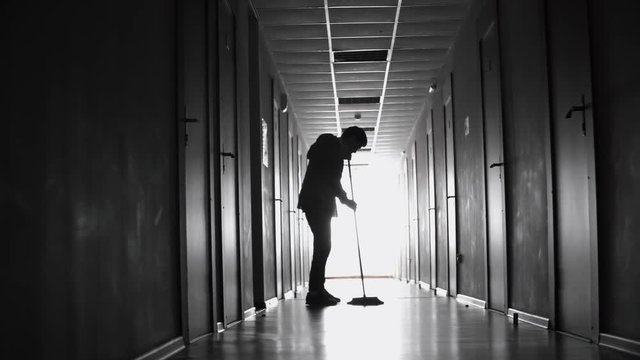 PAN with rear view of silhouette of male janitor sweeping floor in dark corridor 