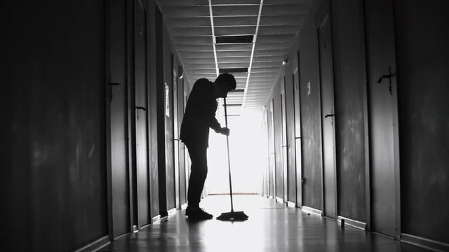 PAN of silhouette of unrecognizable male cleaner sweeping floor with broom in dark hallway 