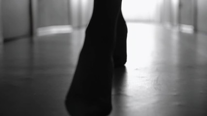 Dolly with low-section of silhouette of female legs in high heels shoes walking along corridor; black and white slow motion shot
