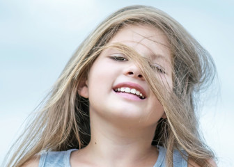 Little beautiful happy girl with hair fluttering in the wind. Portrait of a smiling child on blue sky backgroung