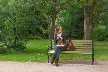 The beautiful woman with a little dog sitting on a bench in the park and waiting for the friend