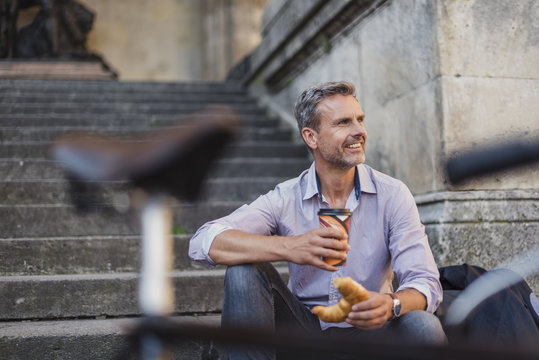 Smiling man sitting on stairs with croissant and takeaway coffee in the city