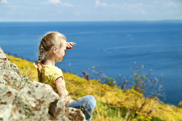 Nice blonde girl with pigtail sitting on the shore of a lake among the yellow flowers and looking into the distance holding her hand above the eyes   
