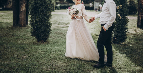 stylish wedding bride and groom walking in sunny park holding hands. modern happy  couple holding...
