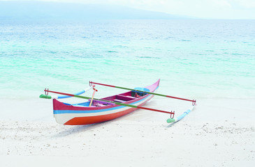 Red white blue colored small empty canoe boat stranded on a tropical white sand beach with blue ocean water on a sunny day in the afternoon.