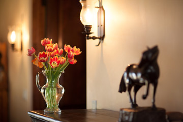 Orange Tulips Bouquet in a Glass Vase next to a Dark Wooden Horse Sculpture on a Rustic Wood Dark Surface, Cream Beige Background, Wall Sconce Lights