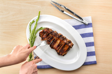 Hands of young woman cutting delicious pork ribs on plate in kitchen