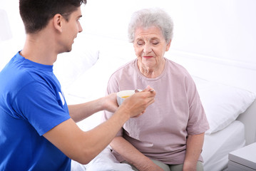 Young male volunteer feeding elderly woman in light room