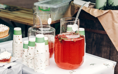 big glass jar with strawberry orange lemonade and paper cups, space for text. street food festival. drink bar at reception, catering outdoors. summer picnic. colorful drinks