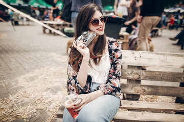 Fototapeta premium stylish hipster woman in sunglasses with red lips holding lemonade and old photo camera. boho girl holding cocktail and smiling at street food festival. summer vacation. space for text