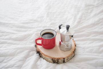 Red enameled metal mug of coffee with steel coffee maker on wooden tray on white bed