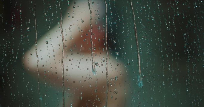 Woman Showering Focus on Water Drops on Door Move Left. a woman from chest up is blurred showering and turning while shot moves left focus on water drops on glass door falling in slow motion
